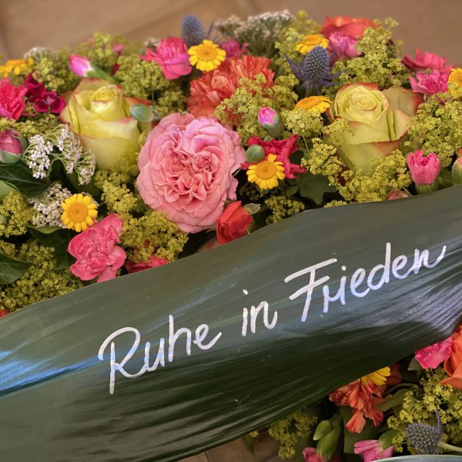 Moderner Trauerkranz ohne Schleife - mit alternative Schleife - Grünes Blatt von Schusterpalme statt Trauerschleife mit Aufschrift "Ruhe In Frieden" ca. 80 cm Durchmesser mit blauen Disteln - Mannstreu, Farbige Freiland Rosen, Frauenmantel, Schafgabe, pinke Nelken, gelbe Färberkamille, Preis ca. 380 Euro